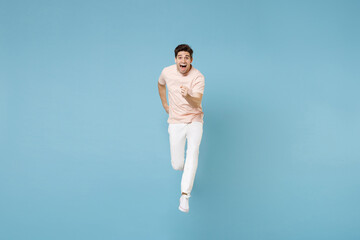 Obraz premium Full length of young excited energetic student man 20s in beige t-shirt white pants do winner gesture clenching fists in air jumping high running isolated on blue color background studio portrait.