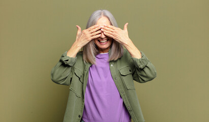 senior woman smiling and feeling happy, covering eyes with both hands and waiting for unbelievable...