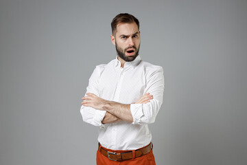 Shocked worried young bearded business man wearing classic white shirt standing hold hands crossed looking camera isolated on grey color background studio portrait. Achievement career wealth concept.