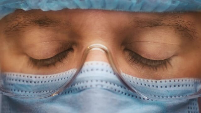 Hospital worker male in medical suit and protective glasses close up. Sad and tired medic opens eyes and looking atraight at camera.