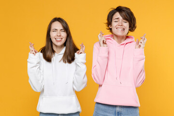 Pleading two young women friends 20s in casual white pink hoodies waiting for special moment keeping fingers crossed making wish eyes closed isolated on bright yellow color background studio portrait.