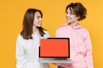 Smiling pretty two young women friends 20s in casual white pink hoodies standing hold laptop pc...