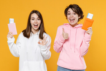 Excited funny two young traveler tourist women friends 20s in casual white pink hoodies hold passport tickets boarding pass showing thumb up okay gesture isolated on yellow background studio portrait.