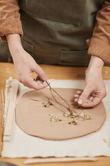Vertical close up of unrecognizable female artisan making plant imprint ceramics on wooden table in pottery workshop, copy space