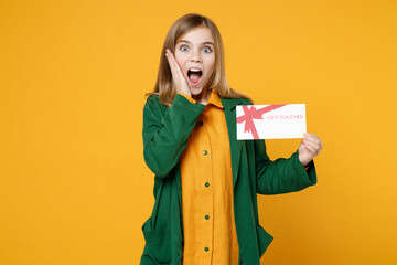 Little blonde shocked happy kid girl 12-13 years old wearing casual clothes green shirt hold gift voucher flyer mock up hold face isolated on yellow background children Childhood lifestyle concept.