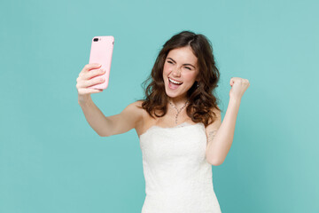 Happy bride young woman 20s in white wedding dress doing selfie shot on mobile phone doing winner gesture isolated on blue turquoise background studio portrait. Ceremony celebration party concept.