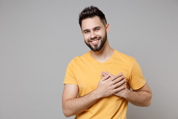 Young caucasian kind good soft-hearted smiling bearded attractive man 20s in casual yellow basic t-shirt put folded hands on heart isolated on grey background studio portrait People lifestyle concept