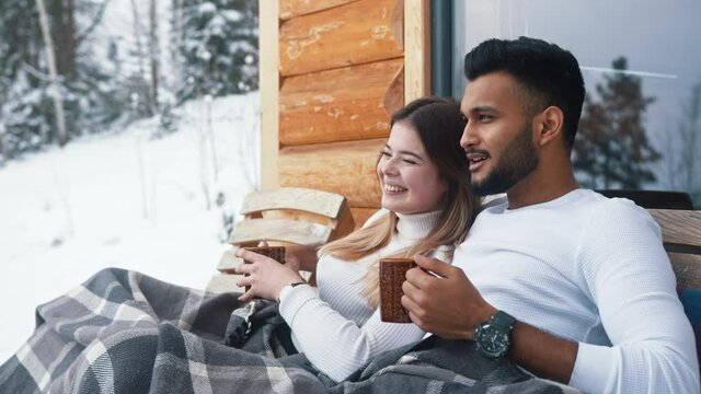 Happy young couple hugging covered with blanket on the outdoors bench. Drinking hot beverage and enjoying snow. High quality 4k footage