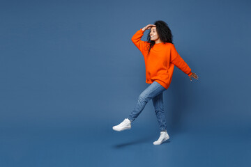 Full length side view of cheerful young african american woman in casual bright orange sweatshirt holding hand at forehead looking far away distance isolated on blue color background studio portrait.