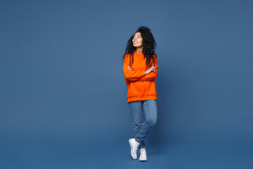 Full length of attractive smiling young african american woman in casual basic bright orange sweatshirt standing holding hands crossed looking aside isolated on blue color background studio portrait.