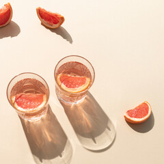 Two glasses of drink with slices of fresh grapefruit on bright beige background. Creative minimal summer concept. Sunny day shadows.