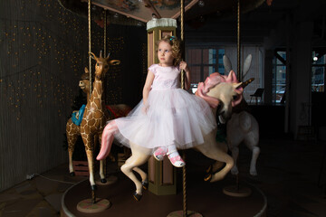 Fototapeta na wymiar a little girl in a pink dress and a crown on a carousel with a white pony