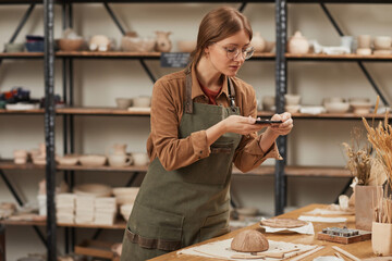 Portrait of young woman taking smartphone photo of raw ceramic bowl in pottery workshop to post on social media, small business concept, copy space