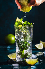 Mojito cocktail, alcohol long drink making. Mint, lime, ice, white rum on blue background. Bartender squeezes lime juice into glass