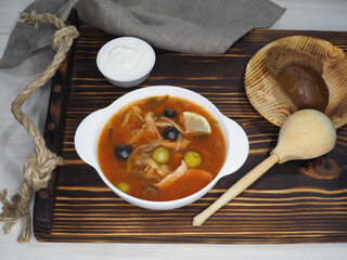 Tomato soup solyanka with olives and sausages, sour cream, bread, wooden spoon on a wooden tray, top view, flat layout. Traditional Russian cuisine, hot dish with meat broth and vegetables for lunch