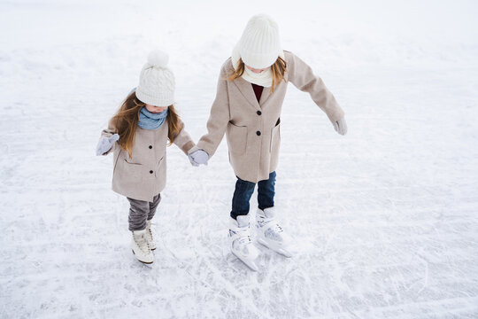 Beautiful little girls, sisters learn to skate on ice skating rink in park. Fall and have fun. Stylish looks, warm woolen coats, white hats, scarves, snoods. Winter family activities, games outdoors