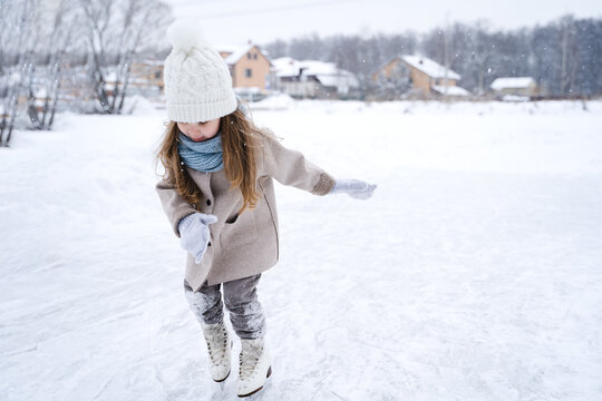 Beautiful little cute girl learn to skate on ice skating rink in park. Fall down and have fun. Stylish look, warm woolen coat, white hat, scarf, snood. Winter family activities, sport, games outdoors