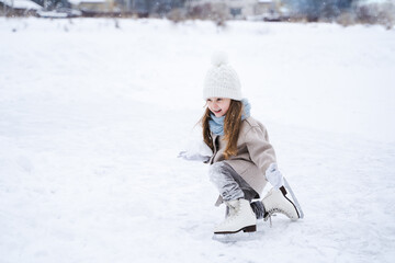Fototapeta na wymiar Beautiful little cute girl learn to skate on ice skating rink in park. Fall down and have fun. Stylish look, warm woolen coat, white hat, scarf, snood. Winter family activities, sport, games outdoors