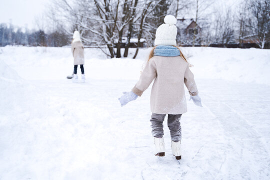 Beautiful little girls, sisters learn to skate on ice skating rink in park. Fall and have fun. Stylish looks, warm woolen coats, white hats, scarves, snoods. Winter family activities, games outdoors