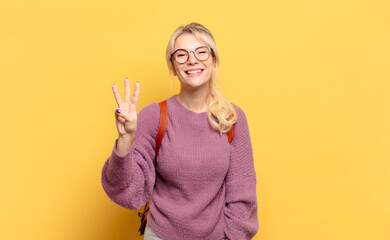 blonde woman smiling and looking friendly, showing number three or third with hand forward,...