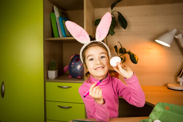 A girl in the ears of an Easter bunny paints eggs with a felt-tip pen in the home interior. Crafts, preparation for a religious holiday, a tray with eggs, hare ears made of plasticine