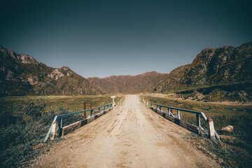 Wide-angle mountain scenery with an old wooden bridge and an earth road stretching into the distance to the ridge in the background, with meadows overgrown with native grasses on the sides, Altai