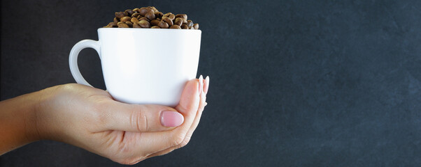 A female hand holds a white cup with coffee beans. Gray stone background.