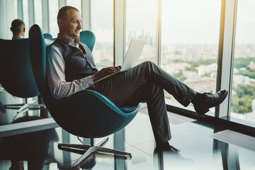 A cheerful adult successful man entrepreneur having a video group call with his partners via the laptop while sitting on a teal armchair in front of an office window with an urban cityscape outside