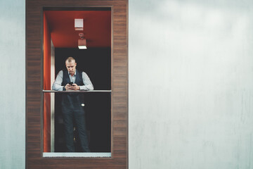 A serious mature businessman in a plaid vest is typing a message via the cellphone while leaning against the chrome railing of a balcony indoors of a modern office with a copy space place on the right