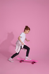 cute little child girl in casual clothes riding skateboard against pink background.