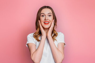 Young caucasian shocked, surprised woman with red lips in a white t-shirt smiling looking at the camera isolated on pink background