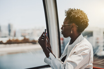 A profile portrait of a young beautiful black tourist woman in sunglasses sitting in the cabin of a ropeway and shooting attractions from the high above using her smartphone or taking a selfie - Powered by Adobe