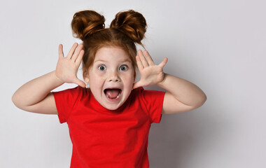 Shocked little girl with freckles and red hair screams loudly with raised hands looking at the...