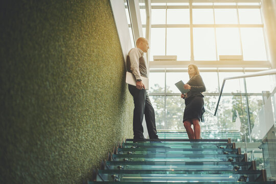 A scene with a glass staircase in the interior of a modern office skyscraper with two business partners having a conversation: businesswoman with a digital tablet and man entrepreneur with a laptop