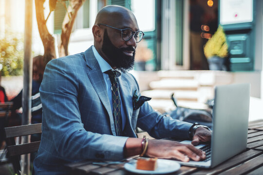 A handsome stylish mature bald black man entrepreneur with a well-groomed beard, in spectacles, and a fashionable suit is using his laptop while sitting in a street cafe during the coffee break