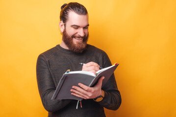 Handsome cheerful young man writing in agenda or planner over yellow background.