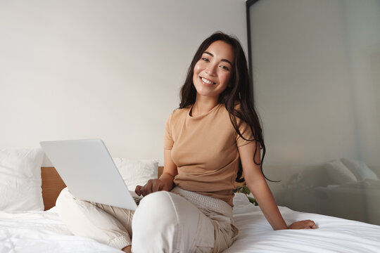 Happy asian woman sitting on her bed with laptop and smiling at camera. Girl relaxing at home and surfing the net in computer