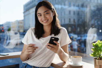 Young asian girl paying for coffee with credit card and smartphone, smiling in cafe near window, drinking cappuccino