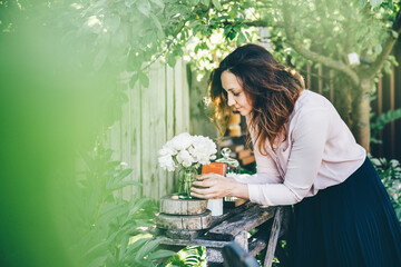 Pretty woman wearing pink blouse puts glass vase with beautiful white peonies near candles and gifts on wooden table near fence in summerhouse yard. - Powered by Adobe