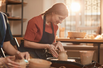 Warm toned portrait of young woman shaping clay on pottery wheel in sunlit workshop and enjoying...