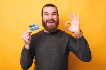 Cheerful young man with beard holding credit card and showing ok gesture over yellow background.