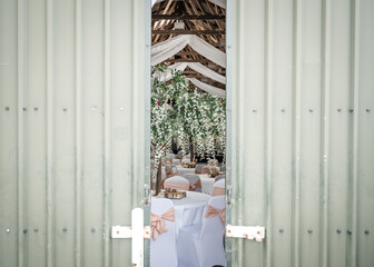 Sneaking a look at wedding reception party laid out behind rustic doors in traditional old converted farm barn with blossom trees white tables and beautiful elegant wooden beam roof ceiling drapes