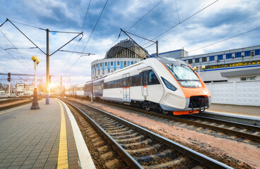 Fototapeta na wymiar High speed train on the railway station at sunset. Industrial landscape with moving modern intercity passenger train on the railway platform, buildings. Railroad in Europe. Commercial transportation