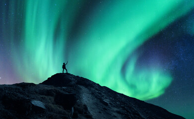 Northern lights and silhouette of standing man with raised up arms on the mountain in Norway. Aurora borealis and happy man. Sky with stars and green polar lights. Night landscape with aurora. Concept - obrazy, fototapety, plakaty
