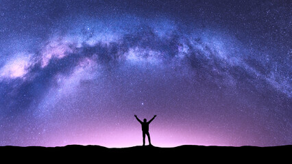 Arched Milky Way and happy man at night. Silhouette of guy with raised up arm on the hill, purple...