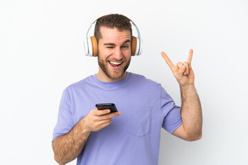 Young handsome caucasian man isolated on white background listening music with a mobile making rock gesture