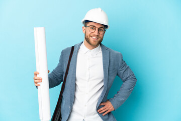 Young architect man with helmet and holding blueprints over isolated background posing with arms at hip and smiling