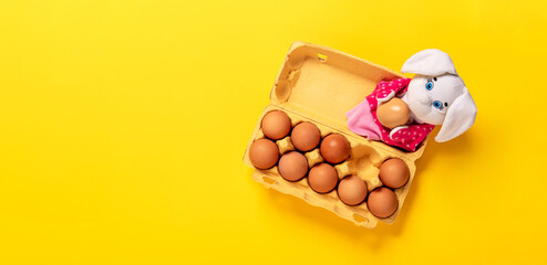 Banner with white Easter bunny laying in the packing with ten eggs on yellow background with copy space, empty text place. Christian holiday card Springtime. Online course of toy theater, hand puppet