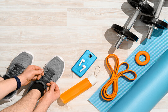 Workout at home concept. Overhead view of a man tying sport shoes with fitness equipment and smartphone on wooden floor. Morning training, sunlight. Place for text, top view.