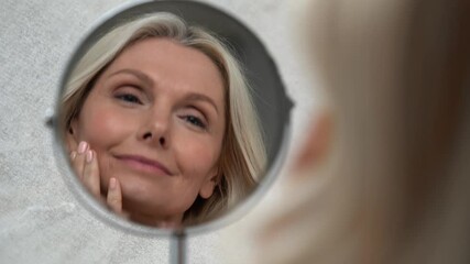 Happy 50s middle aged woman model touching face skin looking in mirror reflection. Smiling mature old lady pampering, healthy moisturized skin care, aging beauty, skincare treatment cosmetics concept. - Powered by Adobe